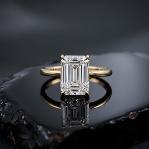 Puede incluir: Un anillo de oro con una gran piedra preciosa transparente de corte esmeralda rectangular, montada en un engaste de cuatro garras. La banda es lisa y pulida, con un acabado metálico reflectante que complementa la piedra facetada central.