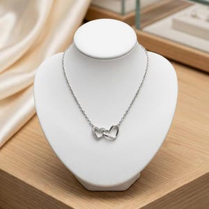 Puede incluir: Un collar de tono plateado con dos colgantes entrelazados en forma de corazón. Un corazón es liso y sencillo, mientras que el otro está adornado con pequeñas piedras claras y brillantes en su borde exterior, suspendido de una fina cadena plateada.