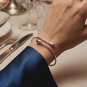 Puede incluir: Una pulsera de plata pulida con un diseño abierto y dos extremos redondeados en forma de gota. El metal tiene una superficie lisa y reflectante que capta la luz, creando una curva elegante y continua alrededor de la muñeca.