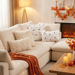 Puede incluir: Un sofá de color crema con dos cojines rectangulares con un patrón de calabazas texturizadas y dos cojines rectangulares con un patrón de fantasmas blancos, junto a una manta de punto naranja quemado sobre el asiento.