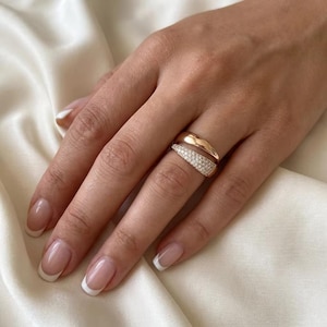 Puede incluir: Un anillo de oro rosa con un diseño de doble banda. Una banda es lisa y pulida, mientras que la otra está incrustada con pequeñas piedras claras y brillantes que cubren la superficie, creando un aspecto texturizado y reflectante.