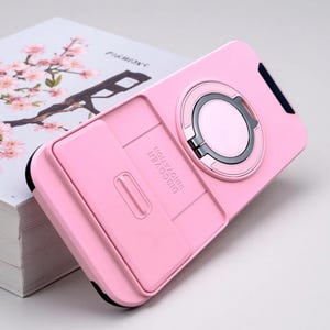 May include: A light pink protective phone case featuring a circular metal ring stand and a sliding camera lens cover. The back of the case includes the embossed text DISCOVER INNOVATION. The design incorporates black accents along the edges.