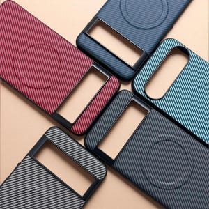 May include: Five smartphone cases featuring a textured carbon fiber pattern in red, dark blue, light blue, black, and gray. Each case includes a black camera bar cutout and a circular magnetic charging ring integrated into the back panel design.