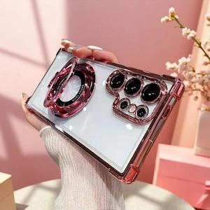 May include: A clear smartphone case with a metallic rose gold frame and reinforced corners. The back features a circular rose gold ring stand with the text KATHY TECH and a raised rose gold camera lens protector with black circular cutouts.