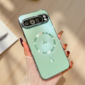 May include: A light mint green smartphone case featuring a metallic camera module cutout at the top and a circular magnetic charging ring design with a central lightning bolt icon printed on the back surface of the protective cover.
