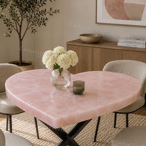 Puede incluir: Una mesa de comedor con forma de corazón, superficie rosa pálido lisa y base de metal negro en forma de X. La mesa está rodeada por cuatro sillas tapizadas con respaldos redondeados y patas de metal negro, formando un conjunto completo.