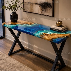 Op de afbeelding: Een rechthoekige consoletafel met een blad van natuurlijk hout en een glanzende blauwe en turquoise harsinleg die stromend water nabootst. De tafel wordt ondersteund door een zwart metalen frame met een X-vormig onderstel aan beide zijden.