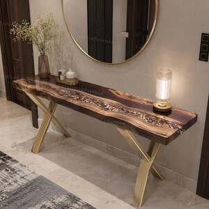 Puede incluir: Una mesa consola rectangular con un tablero de madera oscura que presenta una incrustación central tipo río de pequeñas piedras multicolores incrustadas en resina transparente, sostenida por dos patas de metal dorado en forma de X.