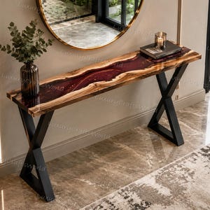 May include: A console table featuring a natural live-edge wood slab with a deep red resin river inlay, supported by black metal X-shaped legs. The surface holds a glass vase with green foliage and a book topped with a small glass candle holder.