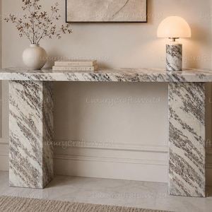 May include: A rectangular console table crafted from white and grey veined marble with two thick slab legs. On the surface sits a white ceramic vase with dried branches, two stacked books titled Kinfolk and Live Beautiful, and a marble lamp.