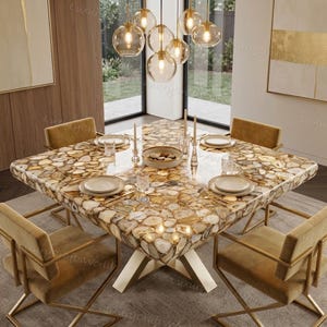 Op de afbeelding: Een vierkante eettafel met een blad van gepolijste, meerkleurige bruine en crèmekleurige agaatplakken. De tafel rust op een goudkleurig metalen onderstel en is omringd door vier bijpassende stoelen met een gouden frame en beige bekleding.