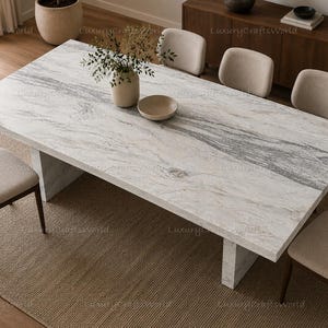 Pode incluir: Uma mesa de jantar retangular com um tampo de mármore branco e cinza e uma base de pedestal espessa. A mesa é cercada por seis cadeiras estofadas com estruturas de madeira escura. Um vaso de cerâmica com folhagem verde fica no centro da mesa.