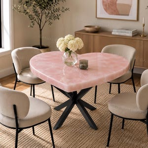 May include: A heart-shaped dining table with a smooth, light pink surface and a black metal base, accompanied by four beige upholstered chairs with black metal legs. A clear glass vase with white flowers sits in the centre of the table.