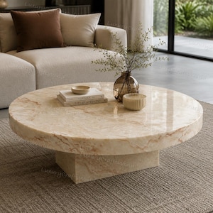 May include: A round, low-profile coffee table crafted from polished beige marble with natural veining. The table features a thick, cylindrical top supported by a rectangular base. On the surface sit two stacked books, a small bowl, and a glass vase.