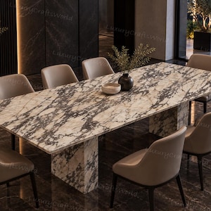 Op de afbeelding: Een rechthoekige eettafel met een wit en zwart marmeren blad en een bijpassende marmeren voet, omringd door acht beige gestoffeerde stoelen met donkere poten. Een kleine kom en een vaas met groen staan op het tafeloppervlak.