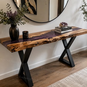 May include: A console table featuring a natural live-edge wood slab with a central translucent purple resin river inlay. The table is supported by two black metal X-shaped legs. A black vase with purple flowers and a black candle sit on the surface.