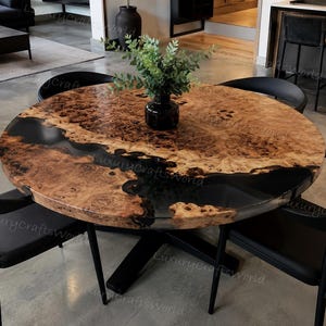 May include: A round dining table featuring a natural wood burl top with a prominent black resin river inlay, supported by a black metal base. The table is surrounded by four black chairs, and a small black vase with green foliage sits in the center.