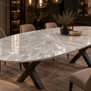 Puede incluir: Una mesa de comedor ovalada con un tablero compuesto por numerosas baldosas rectangulares iridiscentes y claras dispuestas en un patrón de rayos de sol. La mesa se apoya sobre una base de metal negro con dos patas en forma de X.