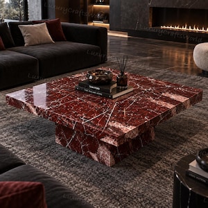 May include: A square coffee table crafted from deep red marble with prominent white veining. The table features a low-profile, block-style base and a polished surface. Resting on top are a dark glass bowl, a reed diffuser, and a book titled Tom Ford.