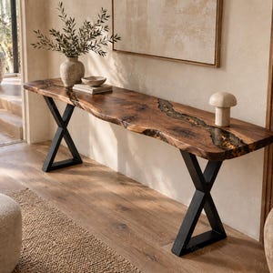 Puede incluir: Una mesa consola larga con un tablero de madera natural de borde vivo y una incrustación central de resina con elementos dorados. La mesa se apoya sobre dos patas de metal negro en forma de X y está colocada contra una pared neutra.