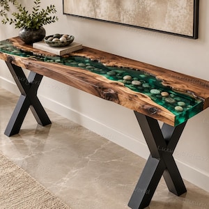 Può includere: Un tavolo console rettangolare con un piano in legno a bordo naturale e un inserto centrale in resina verde traslucida con ciottoli di fiume incorporati, sostenuto da due gambe in metallo nero a forma di X su un pavimento lucido.