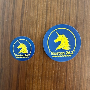 May include: Two circular stickers in different sizes, featuring a blue background with a yellow unicorn silhouette and the text Boston 26.2 printed in yellow at the bottom. The design includes a yellow circular border line.