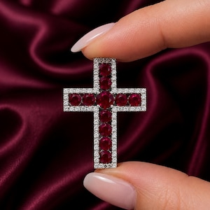 Peut inclure: Un pendentif en forme de croix orné d'une rangée centrale de rubis ronds rouge foncé sertis dans une monture argentée. Les bords extérieurs de la croix sont bordés de petites pierres claires et scintillantes en pavé pour un contraste.
