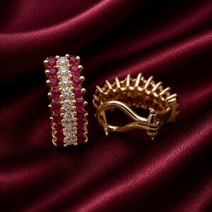 Puede incluir: Un par de pendientes de aro dorados con una fila central de piedras redondas transparentes flanqueada por dos filas de piedras redondas de color rojo intenso. Los pendientes cuentan con un mecanismo de cierre de bisagra y acabado dorado.