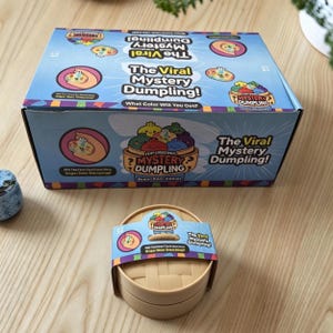 Puede incluir: Una caja de cartón rectangular etiquetada como The Viral Mystery Dumpling con gráficos de dibujos animados coloridos y un recipiente de vapor de plástico beige redondo a juego con una banda de papel con la misma marca e ilustraciones.