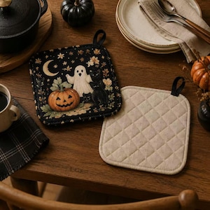 May include: Two square pot holders with black fabric loops. One features a black background with a white ghost, a jack-o'-lantern, black cats, a crescent moon, and stars. The other is a plain, quilted, off-white fabric with a diamond pattern.