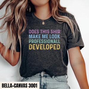 Op de afbeelding: Een donkergrijs gemêleerd T-shirt met korte mouwen en de tekst "DOES THIS SHIR MAKE ME LOOK PROFESSIONALL DEVELOPED" in gestapelde regels in paars, blauw, groen en geel met een verweerd en versleten textuureffect op de print.