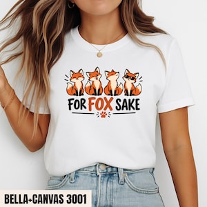 Puede incluir: Una camiseta blanca de manga corta con un gráfico de cuatro zorros de dibujos animados en naranja y negro. El texto "FOR FOX SAKE" está impreso en negro y naranja debajo de los zorros, acompañado de un pequeño icono de huella centrado.