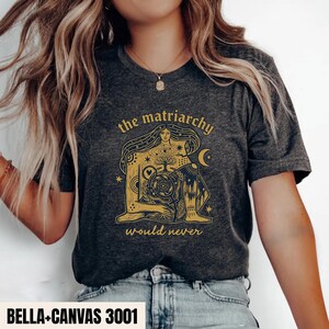 May include: A dark heather gray short-sleeve t-shirt featuring a gold graphic print of a seated figure with long hair, a tree, and celestial elements. The text "the matriarchy" appears above the graphic and "would never" appears below it.