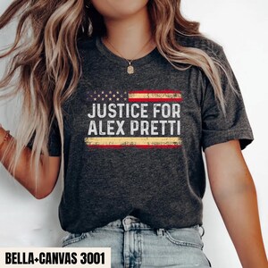 Puede incluir: Una camiseta de manga corta en color gris jaspeado oscuro con un gráfico desgastado de la bandera estadounidense y el texto blanco "JUSTICE FOR ALEX PRETTI" centrado. La prenda tiene cuello redondo y un corte estándar.