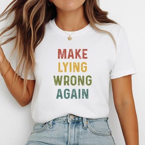 Może przedstawiać: Biały t-shirt z krótkim rękawem z napisem "MAKE LYING WRONG AGAIN" nadrukowanym czcionką w stylu vintage. Słowo "MAKE" jest czerwone, "LYING" żółte, "WRONG" oliwkowozielone, a "AGAIN" morskie, umieszczone centralnie na klatce piersiowej.