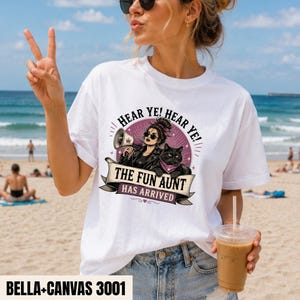 May include: A white short-sleeve t-shirt featuring a graphic print of a person with sunglasses holding a megaphone next to a black cat. The text reads HEAR YE! HEAR YE! THE FUN AUNT HAS ARRIVED. The design includes a purple circular background.