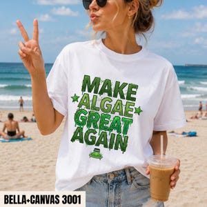 以下が含まれることがあります： 「MAKE ALGAE GREAT AGAIN」という緑色のグラフィックプリントとペトリ皿の小さなイラストが特徴の白い半袖Tシャツ。Tシャツの隅には「BELLA+CANVAS 3001」というテキストが記載されています。