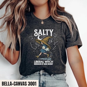 Puede incluir: Una camiseta gris jaspeado oscuro con un gráfico de una persona con un sombrero puntiagudo grande que lleva un frasco de vidrio. El texto dice SALTY LIBERAL WITCH RADICALIZED BY BASIC DECENCY, rodeado de constelaciones de estrellas blancas.
