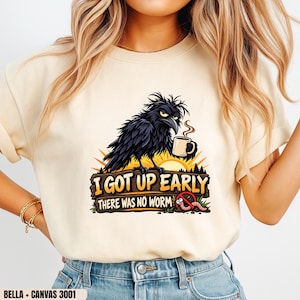 Peut inclure: Un t-shirt à col rond couleur crème avec une illustration d'un corbeau noir tenant une tasse de café. Le texte indique I GOT UP EARLY THERE WAS NO WORM, accompagné d'un dessin d'un ver avec un symbole d'interdiction rouge par-dessus.
