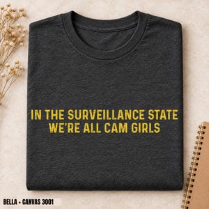 以下が含まれることがあります： ダークチャコールグレーのクルーネックTシャツ。胸の中央に、かすれたサンセリフ体のフォントで「IN THE SURVEILLANCE STATE WE'RE ALL CAM GIRLS」という黄色い文字がプリントされています。半袖で、標準的なフィット感の衣類です。