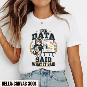 Könnte beinhalten: Ein weißes T-Shirt mit einer Grafik einer getigerten Katze mit Brille und blauem Pullover, die ein Klemmbrett hält und auf ein Diagramm mit der Überschrift Behavior Trends zeigt. Der Text lautet The Data Said What It Said. Label Bella-Canvas 3001.