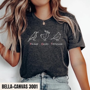 May include: A dark heather gray t-shirt featuring white line art illustrations of three birds labeled Flicker, Dodo, and Titmouse in pink text. The shirt has short sleeves and a crew neckline, with the text BELLA+CANVAS 3001 printed at the bottom.
