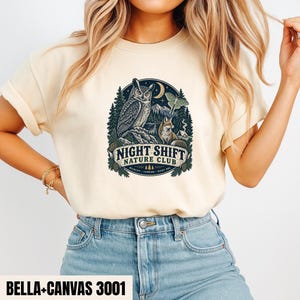 May include: A cream-colored t-shirt featuring a circular graphic with an owl, a fox, a moth, and a crescent moon above a forest landscape. The text NIGHT SHIFT NATURE CLUB is printed in a banner at the bottom of the design, along with smaller text.