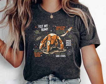 May include: A dark heather gray t-shirt featuring a graphic of an orange crab on a moon surface surrounded by space elements, stars, and planets. The text reads: FIST MY BUMP, USUALLY YOU NOT STUPID, WHY STUPID QUESTION, AMAZE, GRUMPY ANGRY STUPID, GRACE ROCKY SAVE STARS.