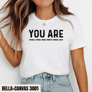 Könnte beinhalten: Ein weißes kurzärmeliges T-Shirt mit dem schwarzen Text YOU ARE, gefolgt von den Wörtern CAPABLE, STRONG, BRAVE, WORTHY, ENOUGH, LOVED. Das T-Shirt hat einen Rundhalsausschnitt und wird mit einer hellblauen Jeans kombiniert, was einen lässigen Stil zeigt.