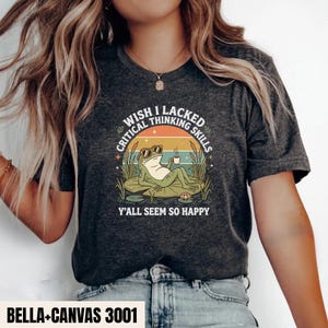 May include: A dark heather grey t-shirt featuring a graphic of a frog wearing sunglasses, lounging on a lily pad while holding a coffee mug. The text reads WISH I LACKED CRITICAL THINKING SKILLS Y'ALL SEEM SO HAPPY, surrounded by cattails and stars.