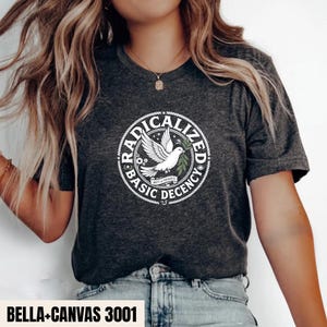 May include: A dark heather grey short-sleeve t-shirt featuring a white circular graphic. The design includes a dove holding an olive branch, surrounded by the text "RADICALIZED BASIC DECENCY" and "UNITY & PROGRESS" in a bold, circular font.