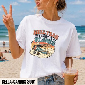 Könnte beinhalten: Ein weißes Kurzarm-T-Shirt mit einer Grafik eines alten Doppeldeckers, der vor einem Sonnenuntergang durch Wolken fliegt. Der Text lautet HELL YEAH I FART IN PLANES in fetter, abgenutzter Schrift. Das Design verwendet orange, blaue und cremefarbene Töne.
