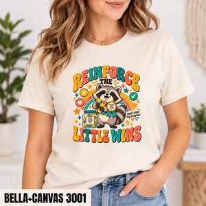 Könnte beinhalten: Ein cremefarbenes T-Shirt mit einer Grafik eines lächelnden Waschbären, der ein goldenes Auszeichnungsband hält. Das Design enthält ein Glas mit bunten Sternen, einen Regenbogen und den Text "REINFORCE THE LITTLE WINS" mit "small steps, big progress".