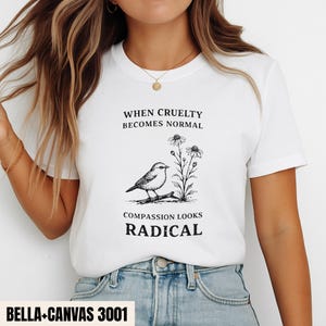 Puede incluir: Una camiseta blanca de manga corta con un gráfico en tinta negra de un pájaro posado en una rama junto a unas flores. El texto dice: WHEN CRUELTY BECOMES NORMAL COMPASSION LOOKS RADICAL. La camiseta se combina con unos vaqueros azul claro.